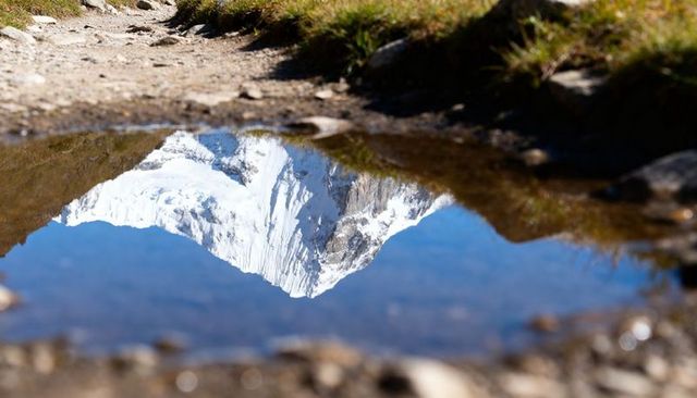 Snowcapped peak reflecting in alpine puddle on rocky trail under clear blue sky