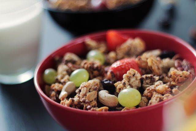 A close-up of a red bowl filled with granola and fresh fruits like berries, grapes, almonds, and cashews. In the background, there is a glass of milk, enhancing the breakfast theme. Suitable for use in diet, health, wellness content, or food blogs and advertisements focusing on nutritious breakfast ideas.