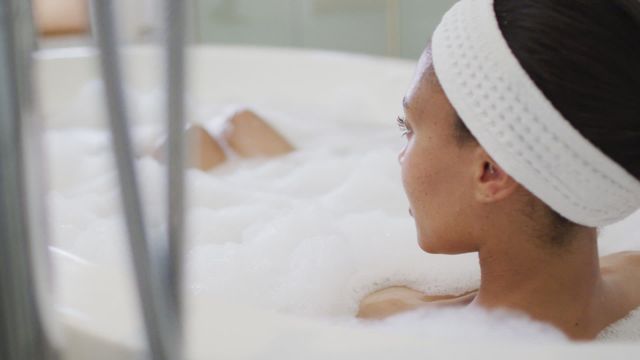 Biracial woman relaxing in a bubble-filled bathtub wearing a headband. Suitable for depicting concepts like self-care, leisure, relaxation, and domestic life. Ideal for wellness blogs, self-care advertisements, and home spa promotions.