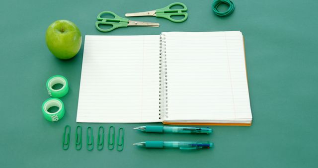 An open notebook lies on a green surface surrounded by school supplies like pens, scissors, and an apple, with copy space. The arrangement suggests a setting prepared for studying or organizing academic work.