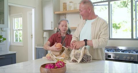 Senior couple unpacking mesh grocery bags at modern kitchen island, preparing fresh fruit