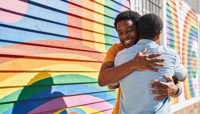 Two men embracing by vibrant rainbow mural celebrating friendship, pride, urban community