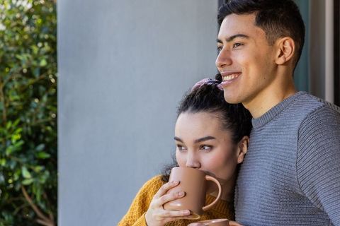 Young couple enjoying coffee on balcony embracing cozy morning together with warm mugs
