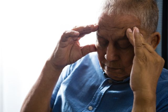 Senior man pressing his head with both hands while sitting by a window in a nursing home. This image can be used to depict themes related to elderly care, health issues, mental health, stress, and aging. It is suitable for articles, blogs, and advertisements focusing on senior healthcare, retirement living, and mental wellness.