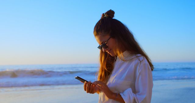 Woman in casual white shirt and sunglasses texting on cellphone while standing by ocean during sunset. Perfect for use in vacation, travel, or tech-related themes, highlighting relaxation and connectivity.