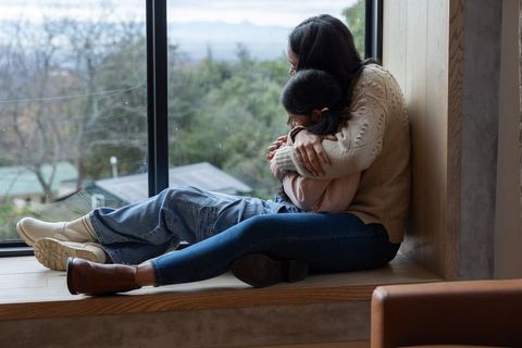 Asian mother hugging daughter on window bench, cozy family bond and nurturing embrace