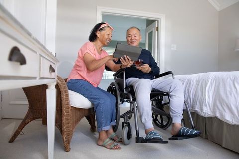 Asian senior couple sharing tablet in bright bedroom, man in wheelchair holding tea