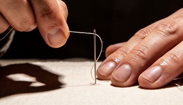 Hands threading needle through fabric closeup showing precise sewing and textile craft
