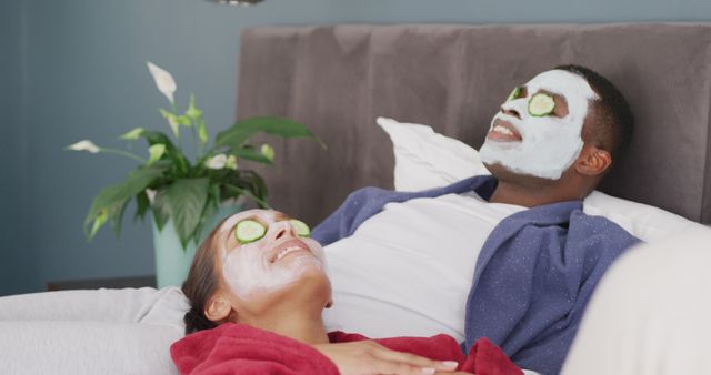 Image of happy diverse couple wearing cleansing face masks and cucumber on eyes lying on bed smiling. Happiness, love, self care, domestic life, and inclusivity concept.
