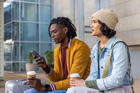 African american travelers waiting outside terminal holding coffee and checking phone