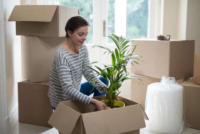 Woman unpacking cardboard boxes in living room, placing houseplant. Ideal for themes of moving, relocation, new beginnings, home organization, and settling into a new home.
