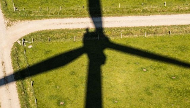 Wind turbine blade shadow casting across green pasture with dirt track and fence posts