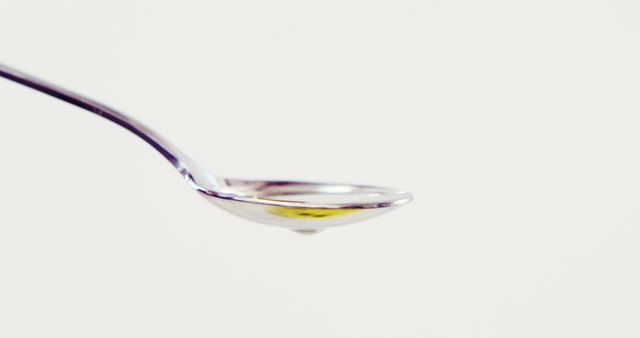 This image features a close-up view of a silver spoon holding a tiny drop of oil against a white background. The simplistic and minimalistic aesthetic makes it perfect for conveying messages related to culinary arts, kitchen tools, cooking ingredients, healthcare, and nutrition. It can be used in articles, blog posts, recipe books, and health forums that focus on cooking tips, ingredient usage, or healthy eating practices.