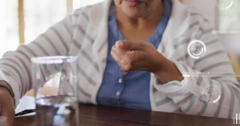 Senior woman reaching for pill with water glass on dining table, home health hud overlay