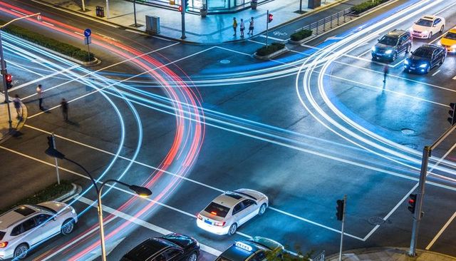 Nighttime urban intersection featuring neon light trails, moving traffic and pedestrians