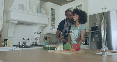 Couple cooking together at kitchen island sharing affectionate embrace, chopping fresh vegetables