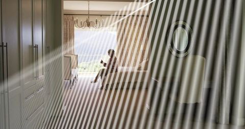 Senior woman sitting on bed edge holding mug, backlit silhouette, balcony view