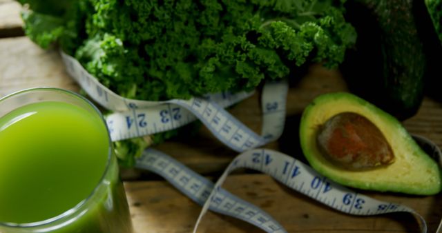 A glass of green juice is placed next to a measuring tape wrapped around fresh vegetables, suggesting a focus on healthy eating and weight management. Avocado and kale signify nutrient-rich food choices for a balanced diet.