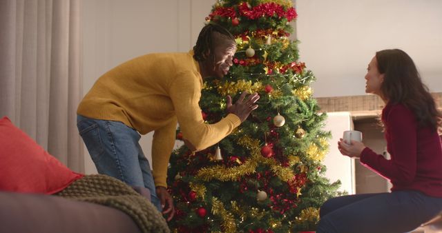 Couple enjoying time together while decorating a Christmas tree at home. Man adding ornaments while woman holds a cup, both smiling and engaging in festive activities. Great for holiday promotions, family lifestyle blogs, and festive advertising.