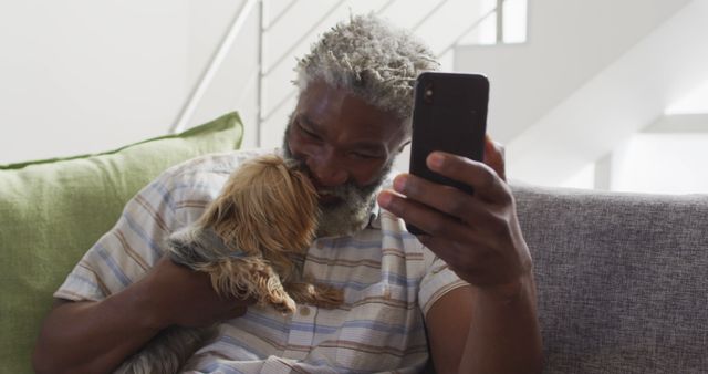 Senior man sitting comfortably on a sofa, holding his small dog close while engaging with his smartphone. Ideal for use in contexts such as senior lifestyle, technology use in everyday life, companionship of pets, or leisure activities at home.