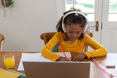 African american girl studying at home with headphones writing in notebook at laptop
