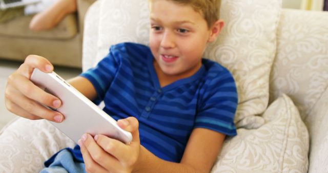 This image shows a young boy in a blue shirt enjoying a game on his tablet while sitting on a comfortable chair. His expression suggests he is thrilled and engaged, making this perfect for educational materials, tech product advertisements, child-friendly apps, or articles on kids and technology use.