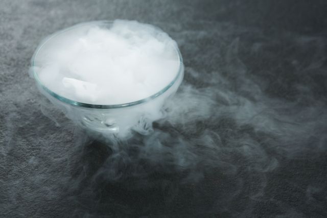 Close-up of dry ice producing smoke in a glass bowl on a dark surface. Ideal for illustrating scientific experiments, chemistry concepts, or creating a mysterious and spooky atmosphere. Useful for educational materials, Halloween-themed designs, or special effects in media projects.