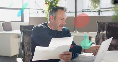 Mature professional analyzing printed charts while pointing at laptop in bright office