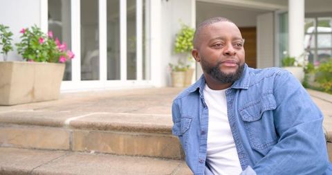 Relaxed african man sitting on porch steps wearing denim jacket and gazing into distance