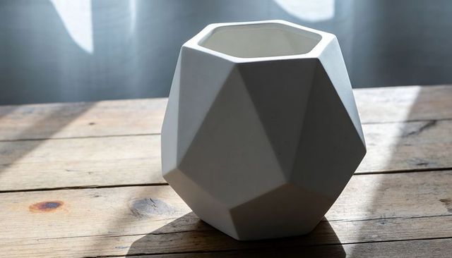 White faceted ceramic vase casting soft daylight shadows on rustic wooden table near sheer curtain