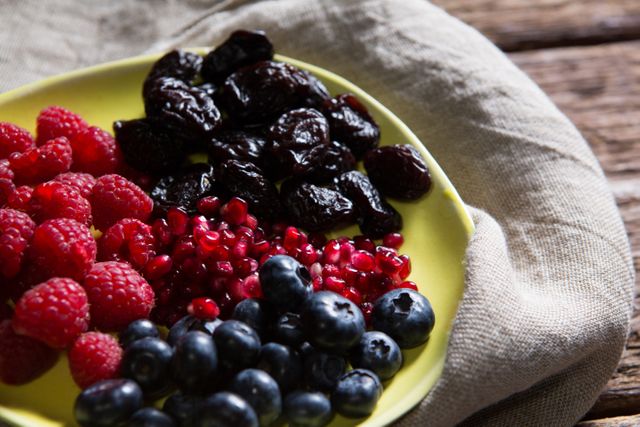 This image showcases a close-up view of a plate filled with fresh raspberries, blueberries, pomegranate seeds, and dried fruits. The vibrant colors and rustic wooden table background make it perfect for use in health and wellness blogs, nutrition articles, and food-related websites. It can also be used in advertisements promoting healthy eating and organic produce.