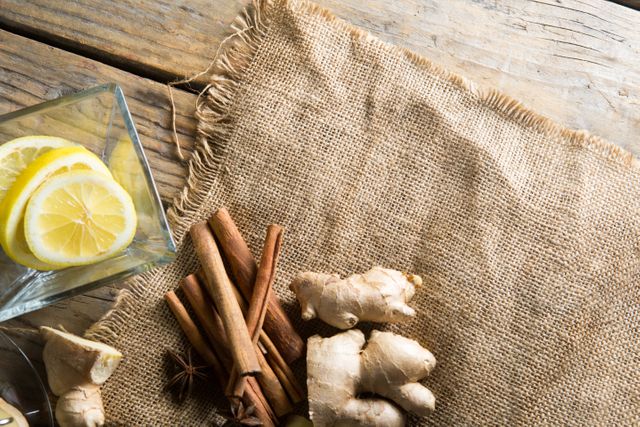 High angle view of fresh ginger and cinnamon sticks placed on burlap fabric with lemon slices in a glass on a wooden table. Ideal for use in food blogs, recipe websites, health and wellness articles, and organic product promotions.