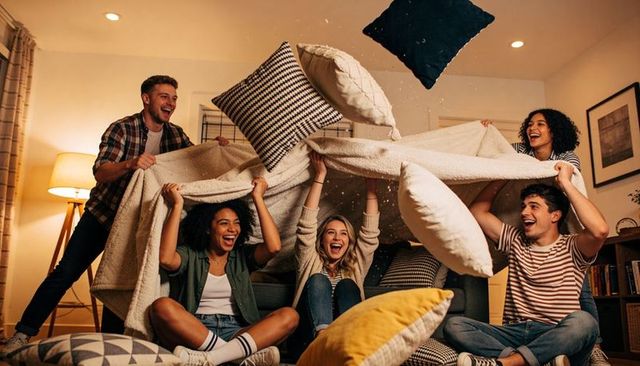 Friends building blanket fort while pillow fighting on living room sofa laughing together