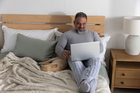 Mature man relaxing and working on laptop in cozy minimalist bedroom with sleeping dog