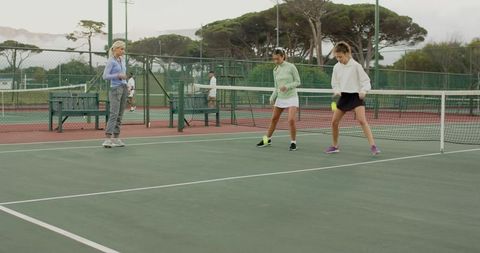Tennis coach guiding women practicing footwork drills on outdoor hardcourt near net