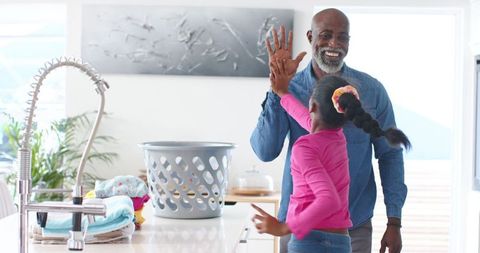 African american grandfather giving high-five to granddaughter doing laundry in modern kitchen