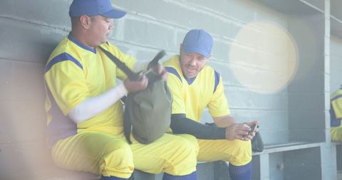 Baseball teammates resting on dugout bench checking smartphone and handling gear bag