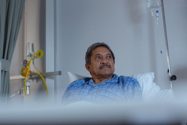 Mature male patient relaxing on a hospital bed in a medical ward. He appears calm and is looking away thoughtfully. The setting includes medical equipment and an IV drip, indicating a healthcare environment. This image can be used for healthcare, medical care, patient recovery, hospital stay, and senior health-related content.