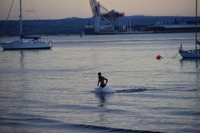 Depicts a person running through shallow water near a harbor at sunset. Silhouette against the backdrop of boats and industrial structures in the distance creates a serene and relaxed atmosphere. Useful for content related to outdoor activities, relaxation by the water, summer evenings, or maritime-themed promotions.