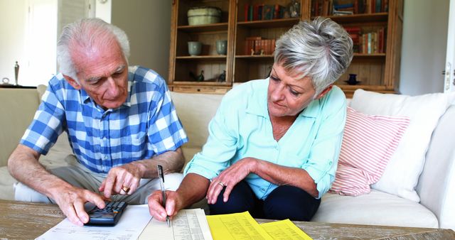 Senior couple sits on a couch in their living room, working on managing their finances. A man holds a calculator and looks focused while a woman writes on a paper. Used for articles or content on elderly financial planning, budget management, retirement strategies, home finance organization.