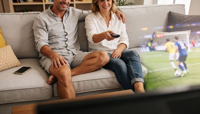 Couple cuddling on sofa holding remote watching live soccer on tv, cozy modern living room
