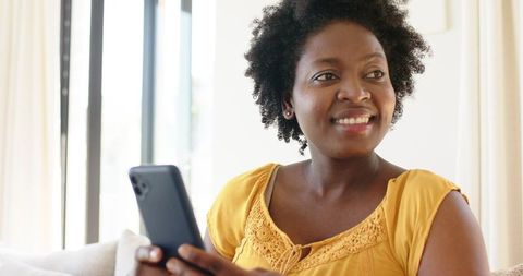 African american woman smiling and holding smartphone on sofa in sunlit living room
