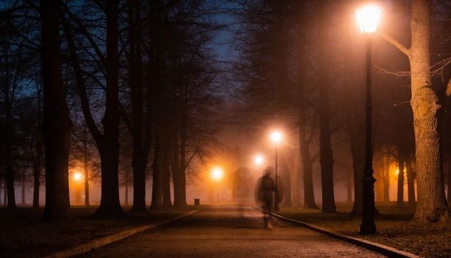 Motion-blurred pedestrian walking through foggy park lit by vintage street lamps at night