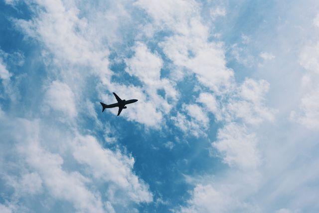 Capturing the silhouette of an airplane flying through a blue sky with scattered clouds. Ideal for travel agencies, aviation-related content, transportation visuals, and themes of freedom and adventure.