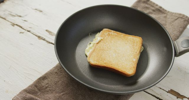 High-angle view of a grilled cheese sandwich cooking in a frying pan placed on a rustic wooden table with a brown towel underneath. This image is perfect for websites or blogs focusing on homemade recipes, comfort foods, easy cooking tutorials, breakfast ideas, and snack suggestions. Ideal for use in food advertisements, culinary magazine articles, and cooking class promotions.