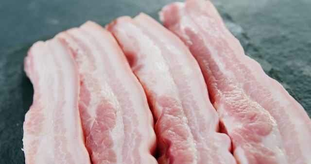 Close-up view of fresh pork belly slices laid out on a dark textured surface. Ideal for use in advertising for butcher shops, cooking blogs, culinary magazines, or food packaging. Perfect for promoting fresh, high-quality meat products.