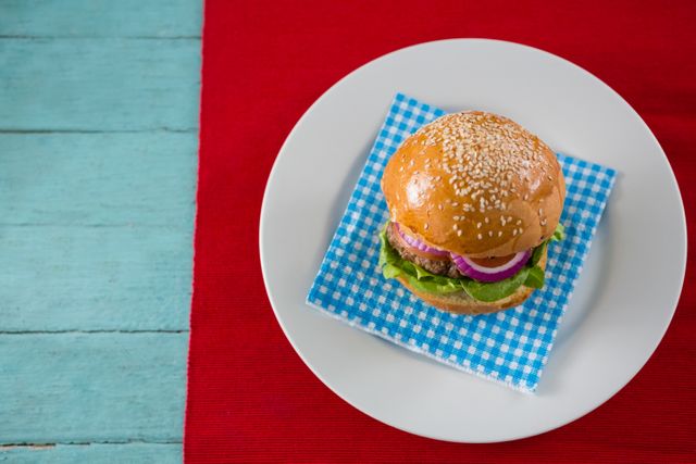 Overhead view of a delicious hamburger served on a blue checkered napkin placed on a white plate. The burger features a sesame seed bun, fresh lettuce, and red onions. The plate is set on a vibrant red tablecloth with a rustic blue wooden table background. Perfect for use in food blogs, restaurant menus, culinary websites, and advertisements promoting gourmet or homemade meals.