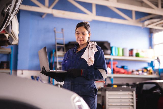 Female car mechanic wearing overalls and face mask using laptop in a garage. Ideal for content related to automotive repair, technology in car maintenance, small business operations, and safety measures during the COVID-19 pandemic.