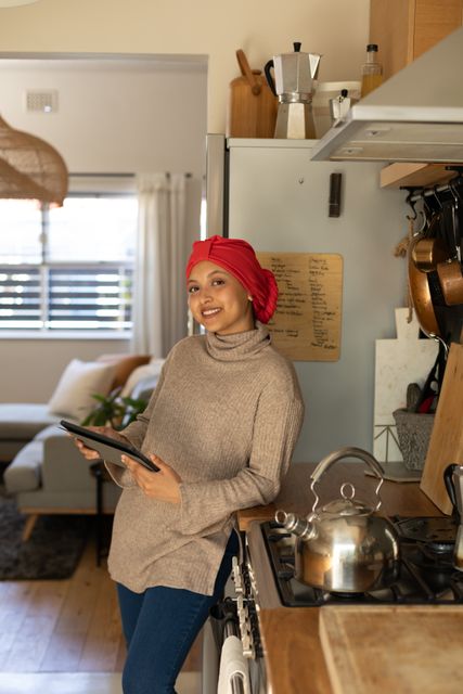 Vertical of smiling biracial woman in hijab using tablet standing in kitchen at home, copy space. Happiness, communication, relaxation, inclusivity and domestic life.