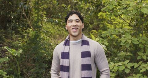 Young man smiling in lush green woodland wearing knit sweater and striped scarf outdoors